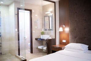 Een moderne hotelkamer met een glazen douchecabine, een houten nachtkastje, een wit bed en een grote spiegel boven de wastafel.