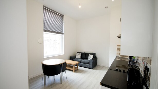 Un espace de vie compact comprenant un coin cuisine, un coin repas avec une table ronde et des chaises, un canapé avec des coussins et une fenêtre avec des stores.