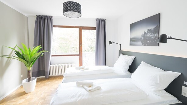 Ein Hotelzimmer mit zwei Betten, einem großen Fenster mit Vorhängen, einem Blumentopf, einer Wandlampe und einem gerahmten Bild.
