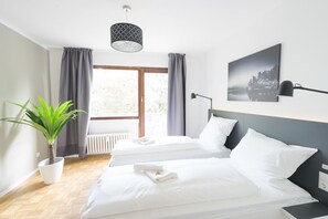 A hotel room with two beds, a large window with curtains, a potted plant, a wall-mounted lamp, and a framed picture. - Glück Auf Appartements Kammerstraße (Duisburg)