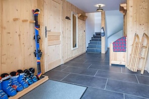 Interno di un rifugio sciistico con parete in legno, porta sci e una scala che conduce a un secondo piano.