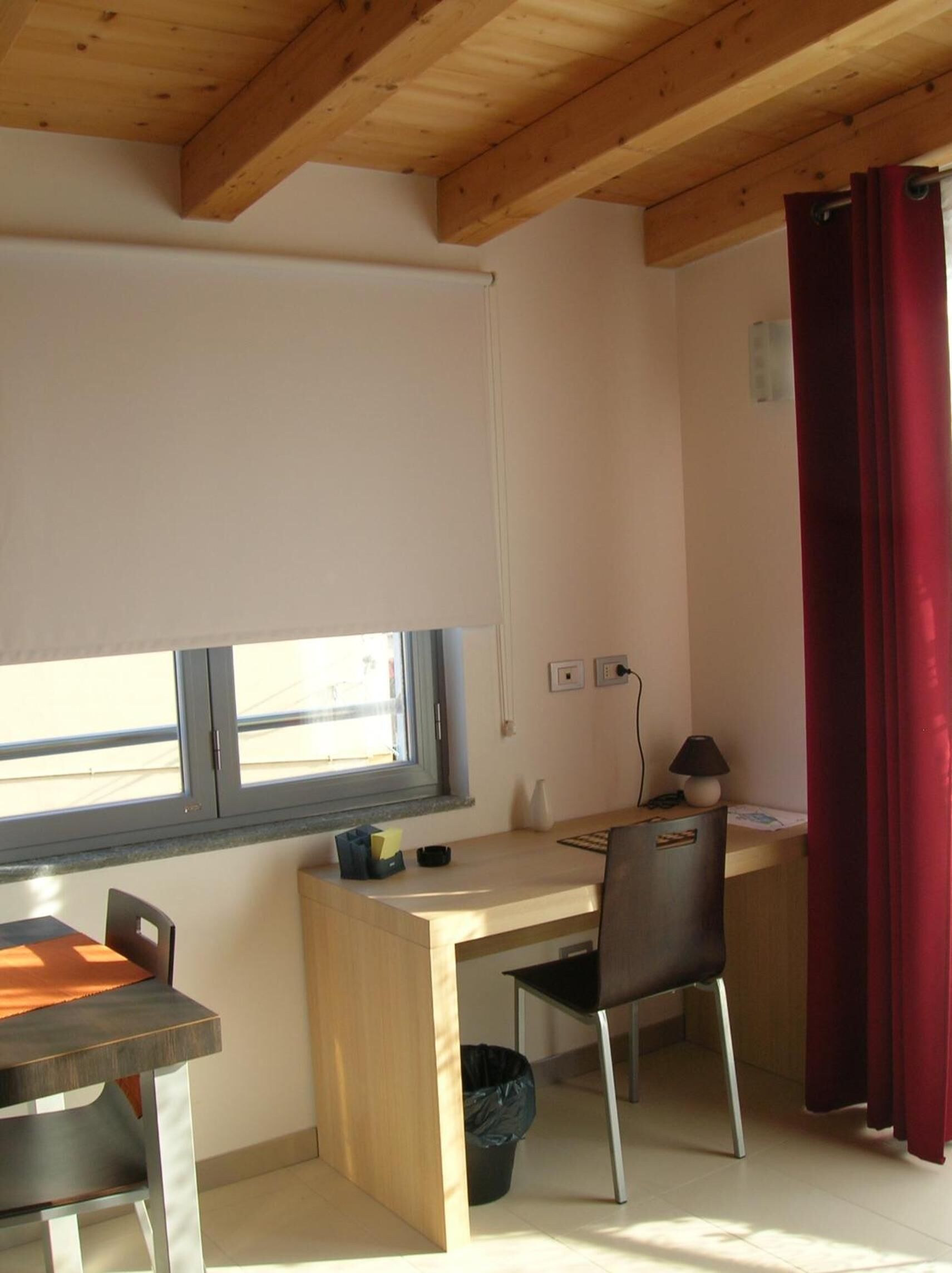 A small, well-lit room with a wooden ceiling, a desk with a chair, a window with blinds, and a red curtain.