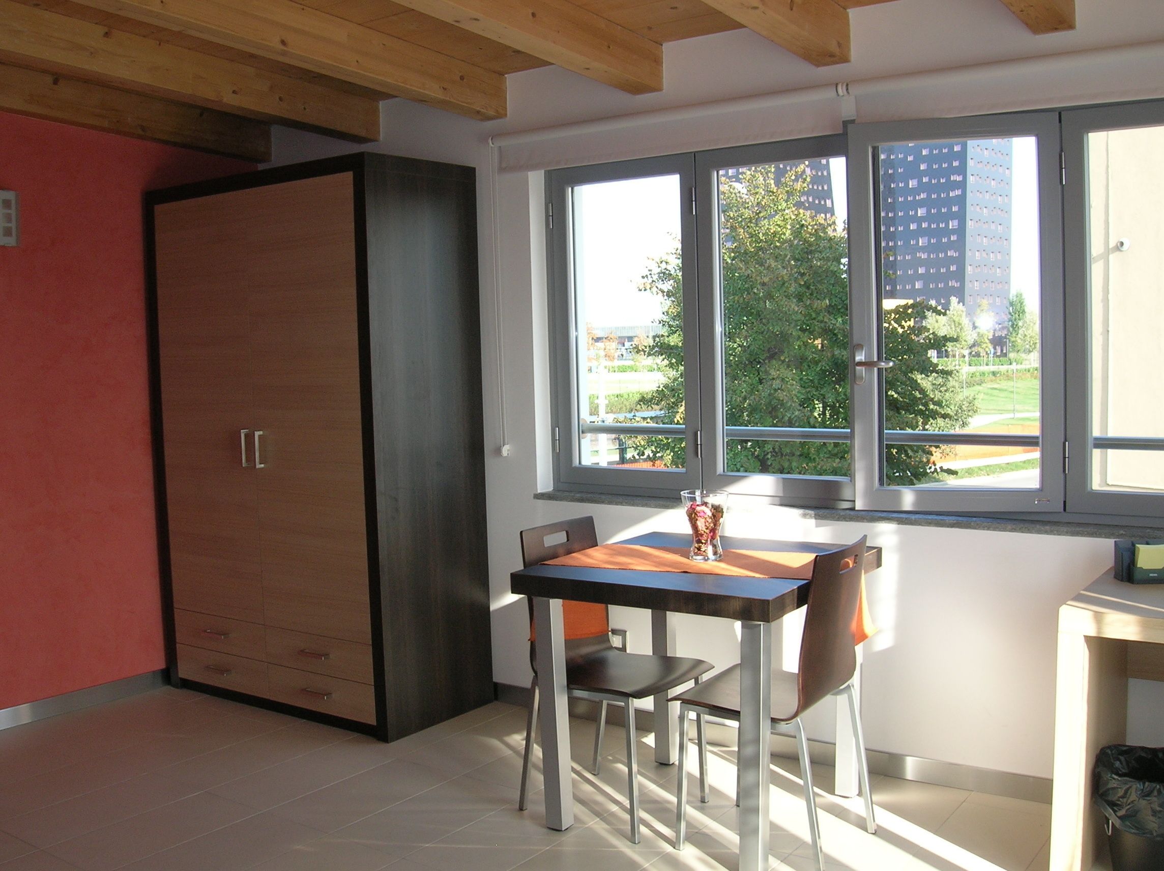 A modern room with a dining table, chairs, a cabinet, and a window with a view of buildings and trees.