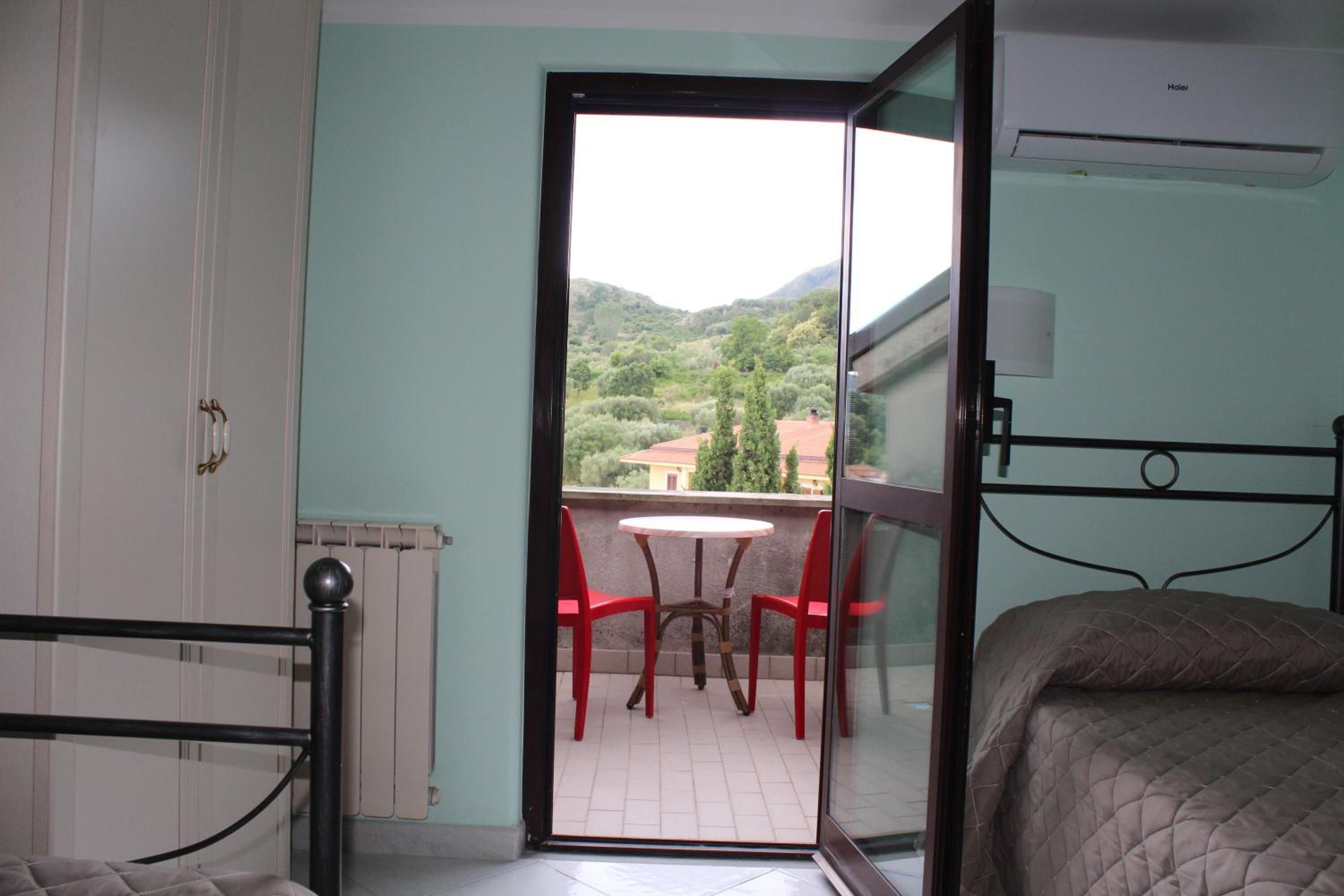 Ein Zimmer mit einem Bett, einem Heizkörper, einem Tisch mit roten Stühlen und Blick auf Grünflächen und Gebäude.