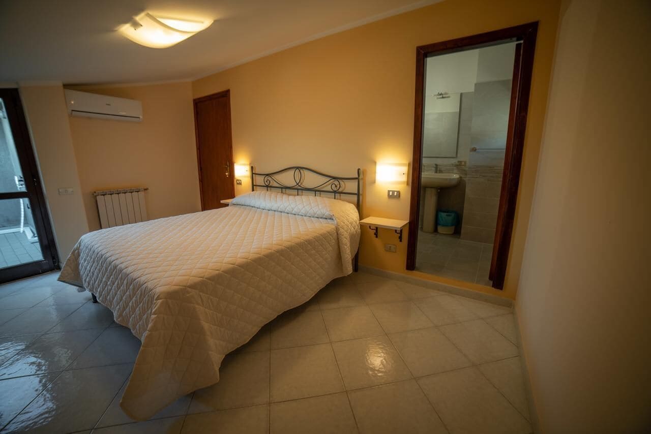 Ein Schlafzimmer mit Bett, Nachttischlampen, einem Heizkörper und einem durch eine offene Tür einsehbaren Badezimmer.