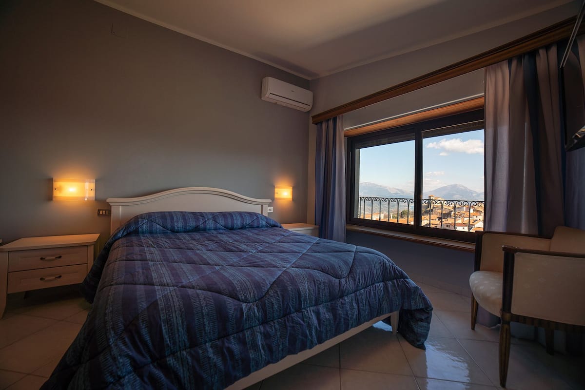 A bedroom with a large bed, bedside tables, an air conditioning unit, a chair, and a window with a view of mountains.