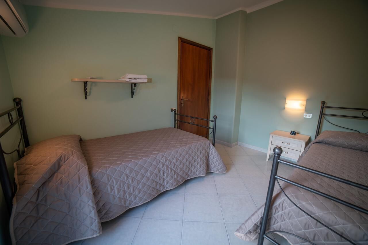 A bedroom with two beds, a wooden door, a shelf with towels, and a bedside table with a lamp.