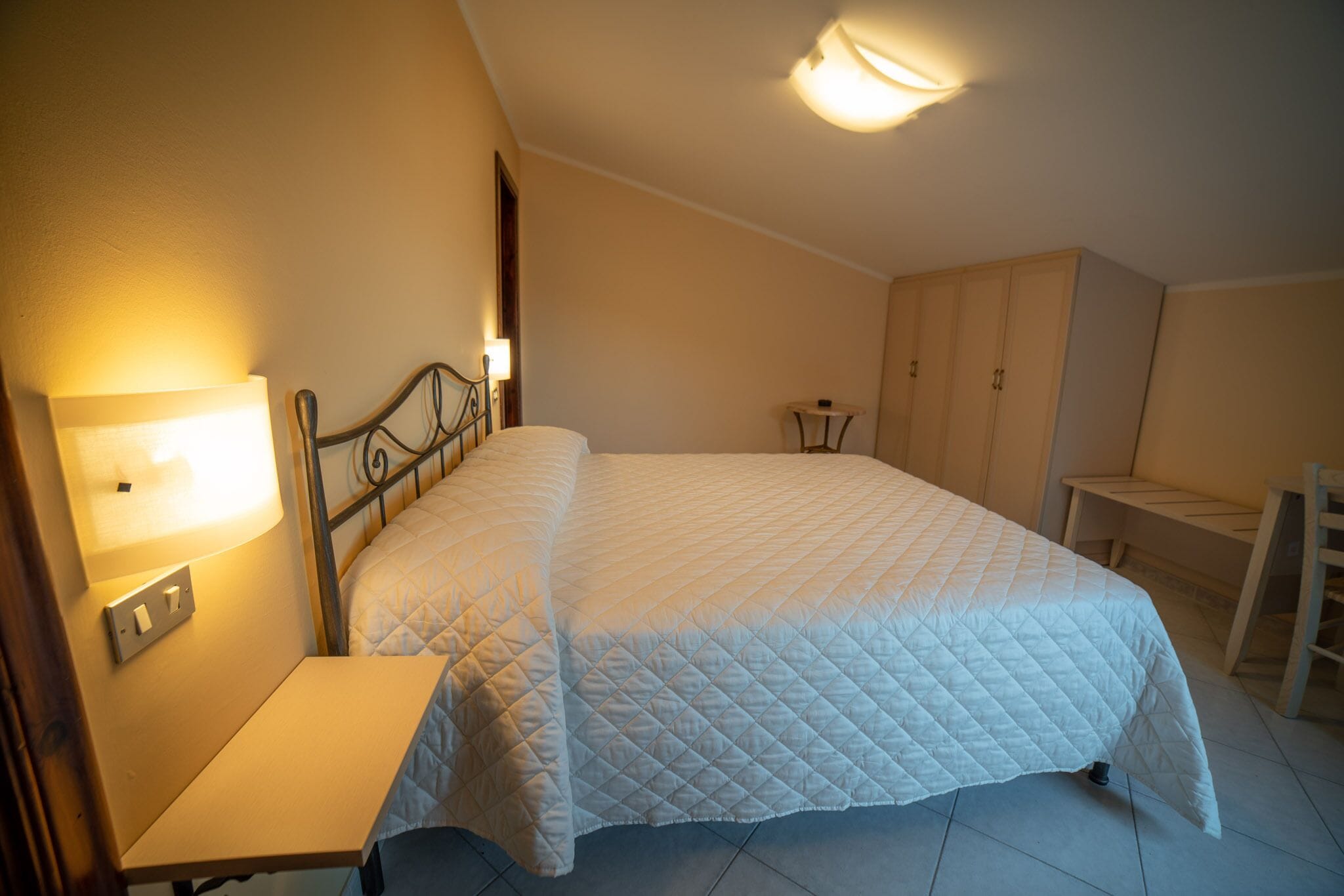 A bedroom with a bed, bedside table, lamp, wardrobe, and a small table.