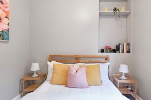 Una cama bien tendida con cabecera de madera, dos mesitas de noche con lámparas y un estante con libros y objetos decorativos.