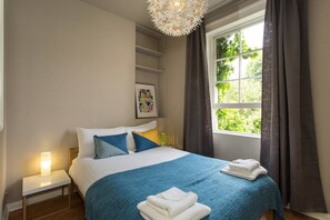 A bedroom with a bed, bedside lamp, window with curtains, and a wall-mounted shelf.
