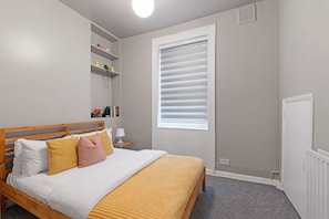 Een slaapkamer met een bed, nachtkastje, lamp, plank met decoratieve objecten, raam met zonwering en een radiator.