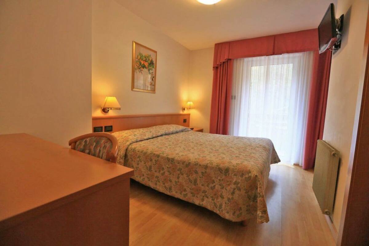 Ein Hotelzimmer mit einem Bett, einem Schreibtisch, zwei Nachttischlampen, einem Bild an der Wand und einem Fenster mit Vorhängen.