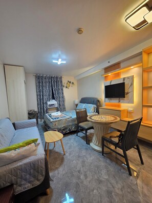 A modern hotel room with a bed, a sofa, a dining table, and a TV mounted on the wall.