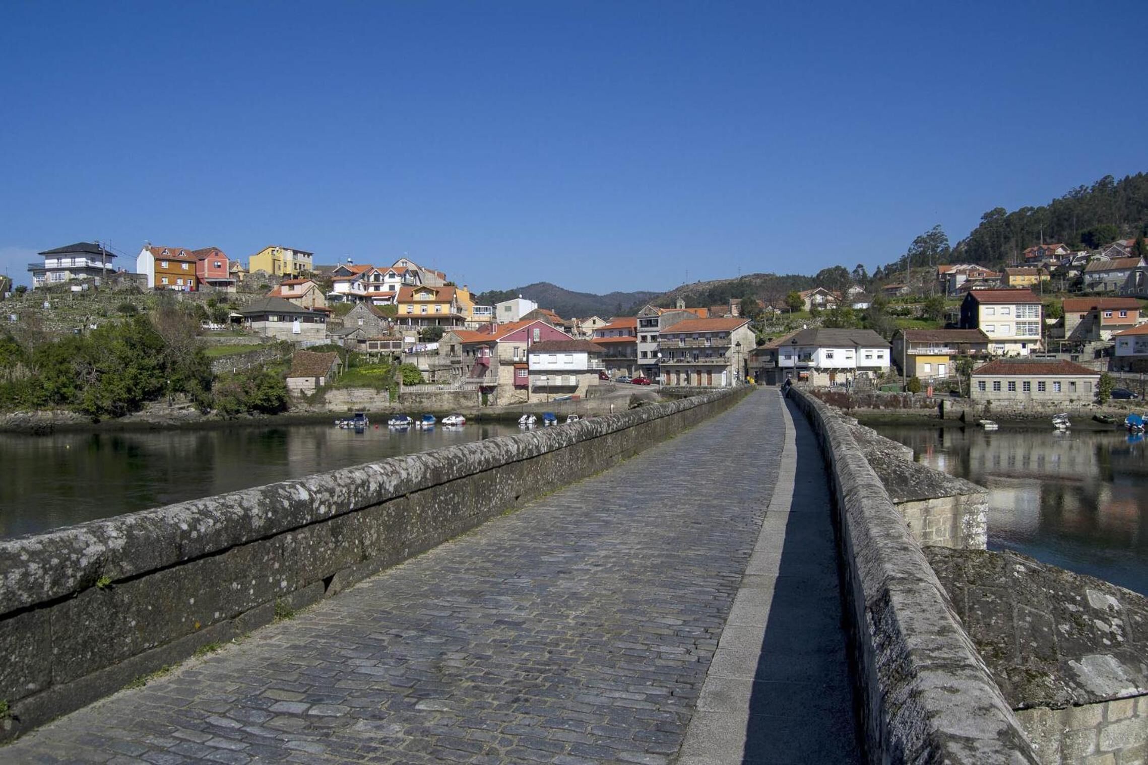 A stone bridge leading to a town with colorful houses and a river.