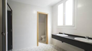 Un baño con piso de baldosas blancas, una puerta de madera, un inodoro blanco y una encimera negra con dos lavamanos.