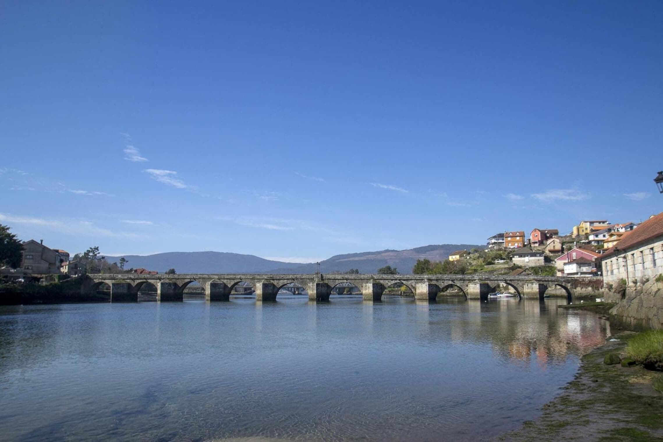 Un puente de piedra sobre un río tranquilo, con casas a la derecha.