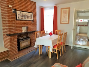 Uma sala de jantar com lareira de tijolos, cadeiras de madeira, mesa com toalha branca e um quadro na parede.