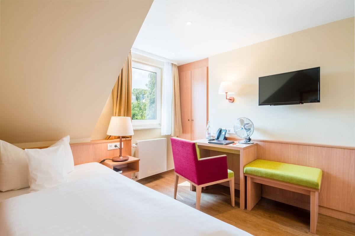 Habitación de hotel con cama, escritorio con silla, televisión y ventana con cortinas.