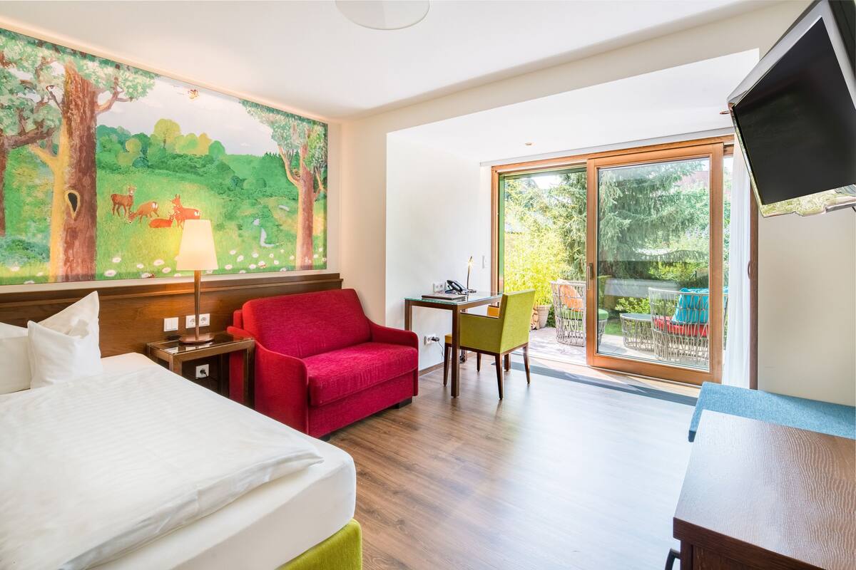 Habitación de hotel con cama, sofá rojo, escritorio con silla y una ventana grande con vista a la vegetación.