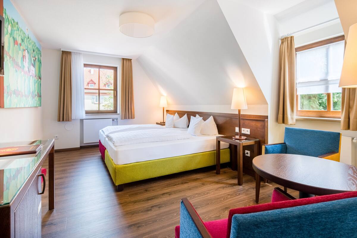 Una habitación de hotel con cama, una mesa, una silla y un ventanal con cortinas.