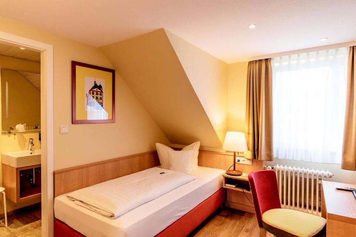 Habitación de hotel con cama, escritorio, silla, radiador, ventana con cortinas e imagen enmarcadas en la pared.