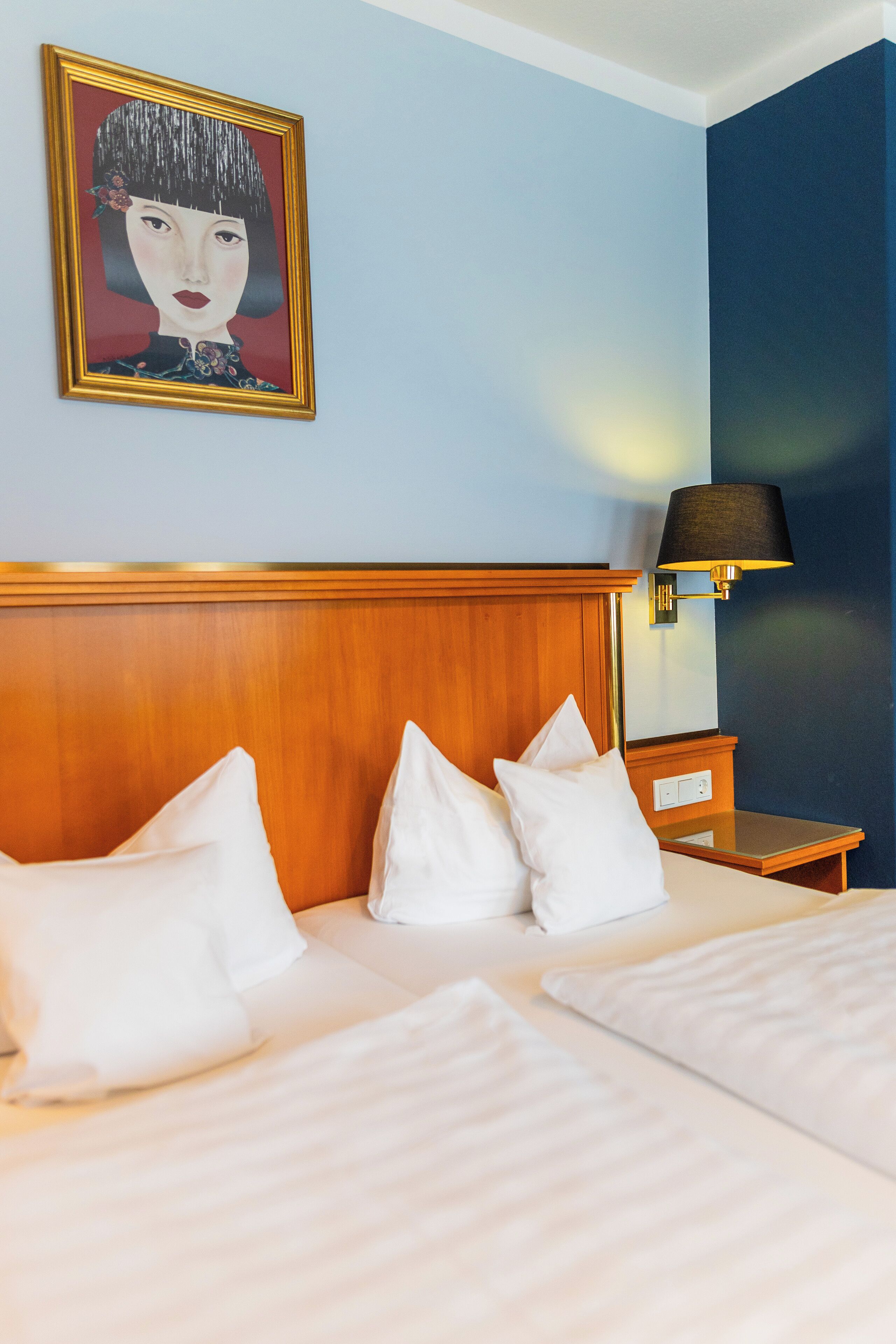 A hotel room with a wooden headboard, two beds with white linens, a nightstand with a lamp, and a framed artwork on the wall.