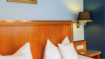 A hotel room with a wooden headboard, two beds with white linens, a nightstand with a lamp, and a framed artwork on the wall.