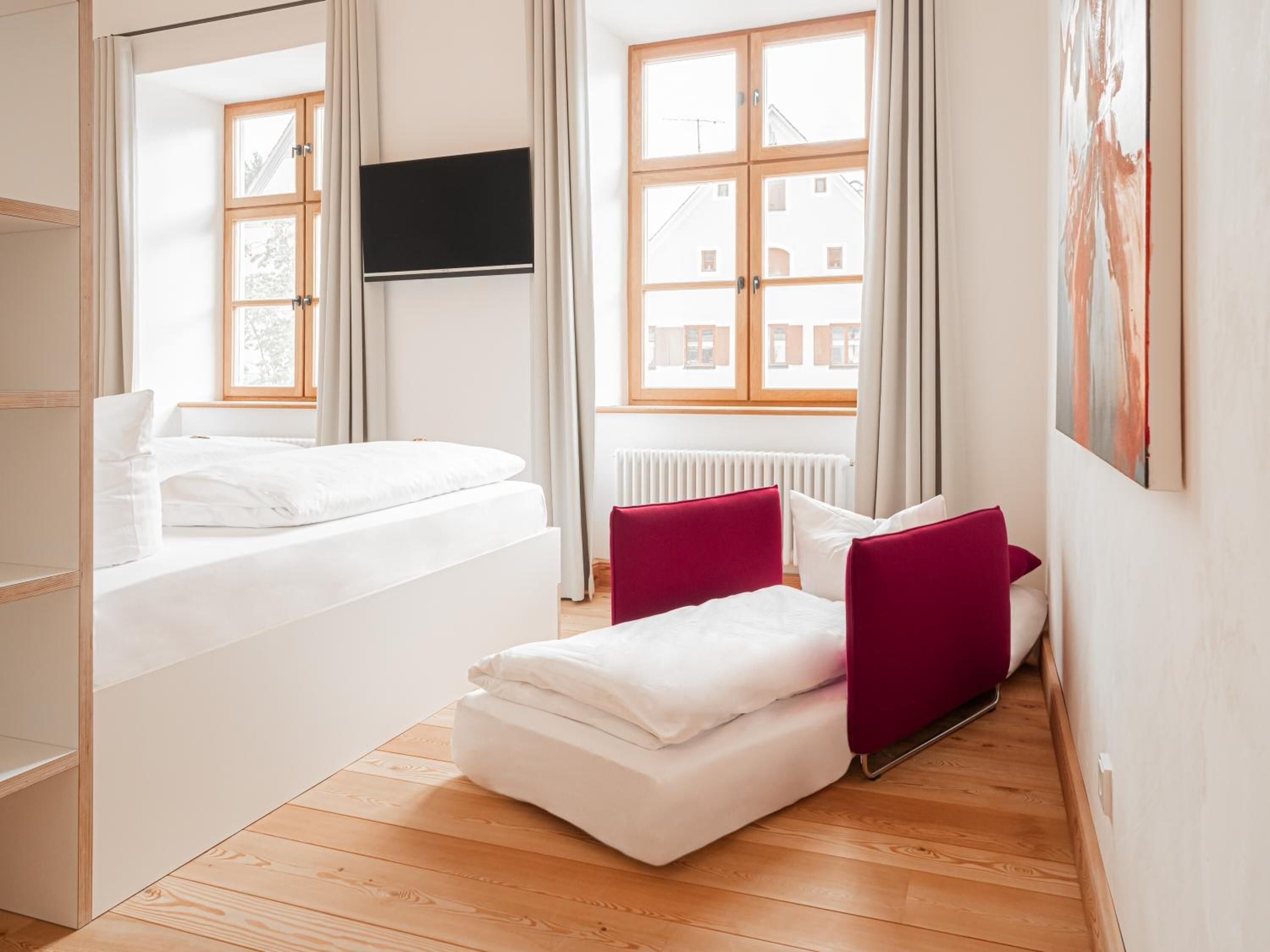 Une chambre d’hôtel moderne avec un lit, un coin salon doté de coussins rouges, un téléviseur à écran plat fixé au mur et de grandes fenêtres avec des rideaux.