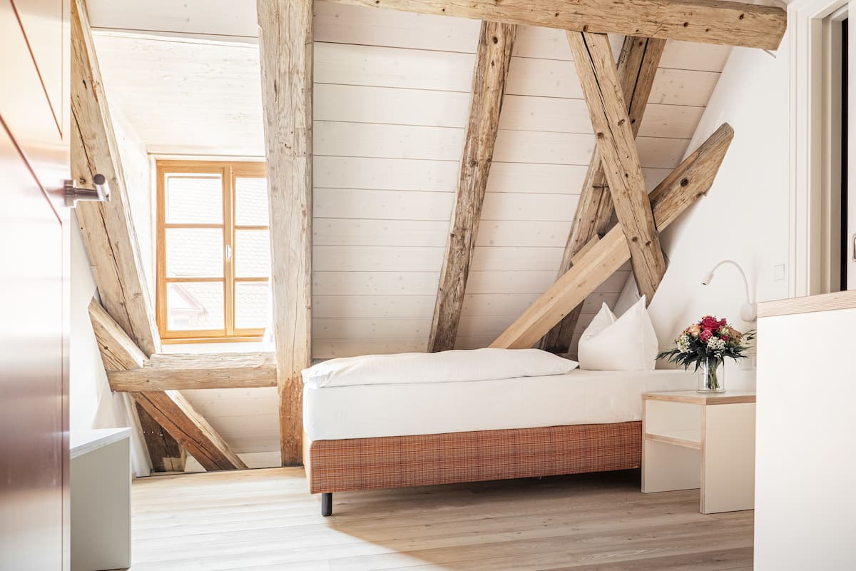 A bedroom with a sloped ceiling, wooden beams, a bed with white bedding, a bedside table with a vase of flowers, and a window with a view of the outside.