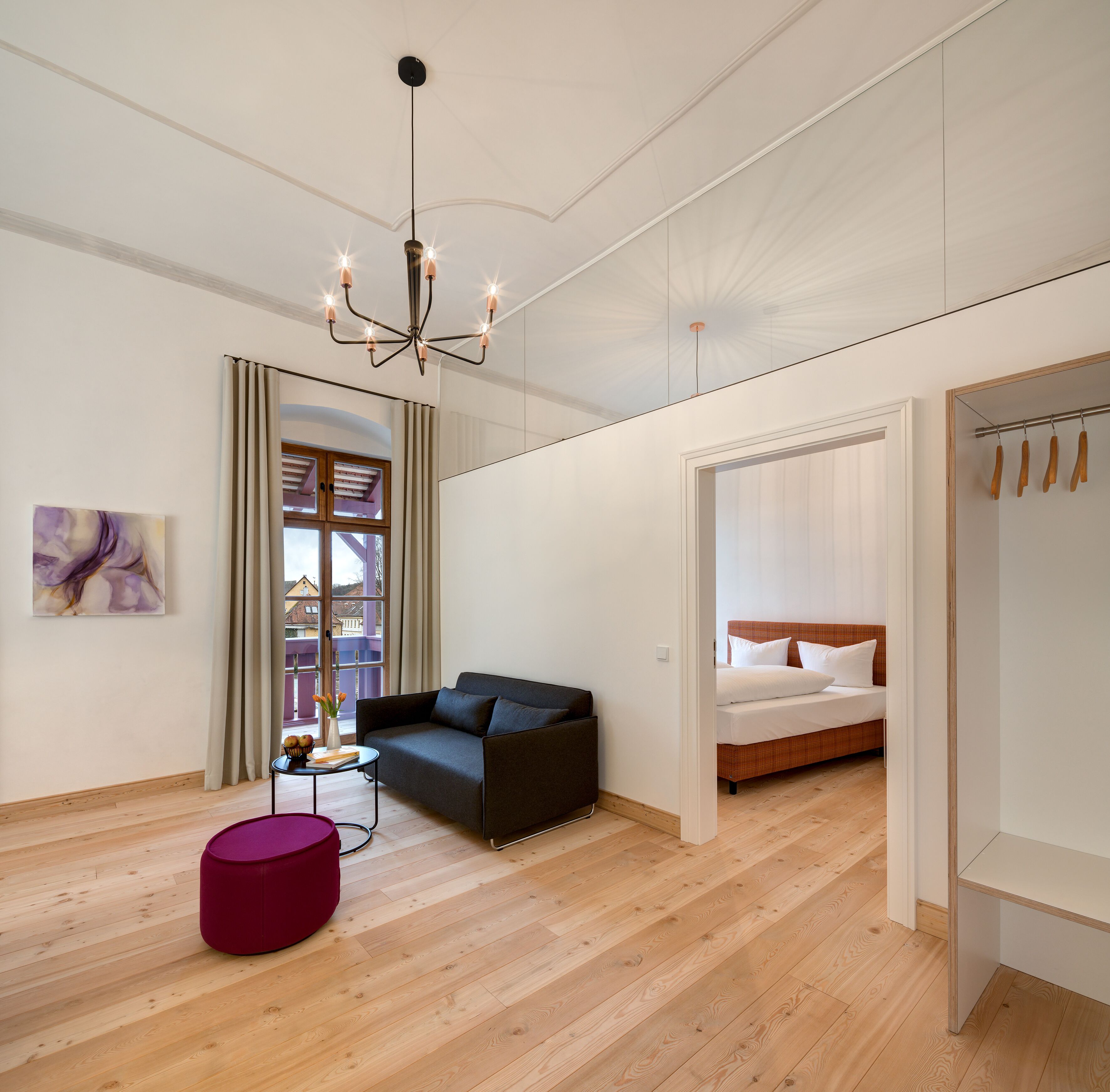 A modern bedroom with a dark sofa, a bed with white bedding, a wooden floor, a pendant light, and a view through a window with curtains.