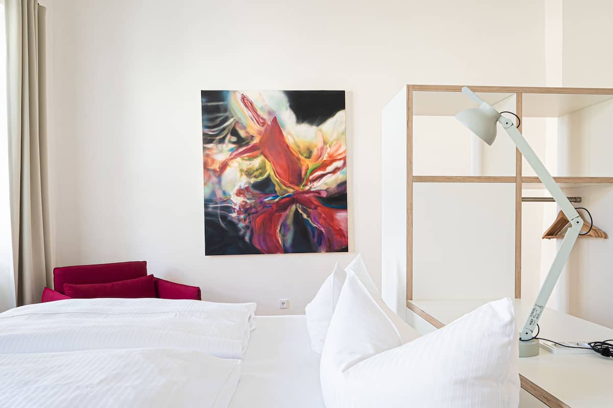 A hotel room with a bed, a red chair, a white lamp, and a colorful abstract painting on the wall.