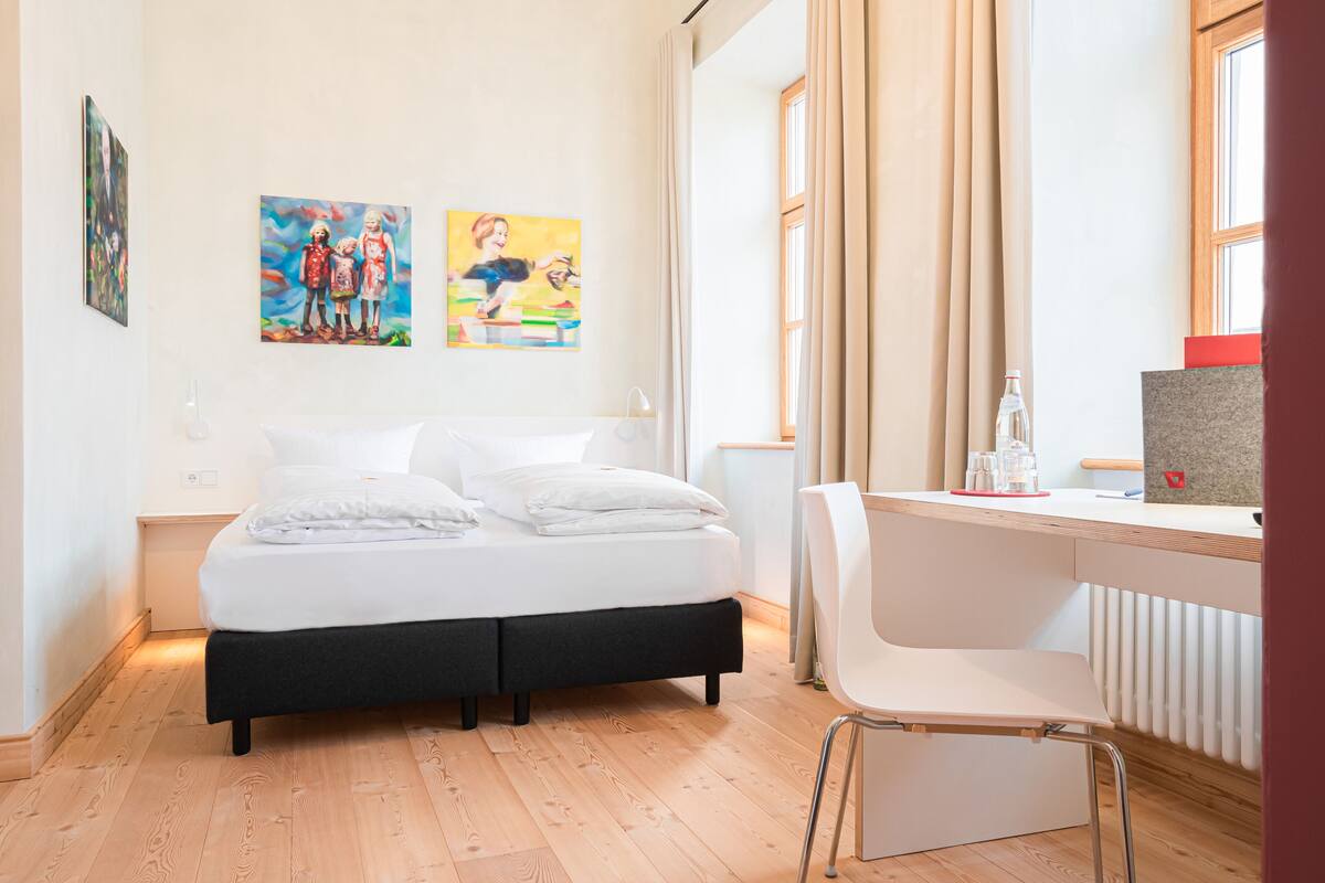 A modern hotel room with a bed, a desk, and two paintings on the wall.