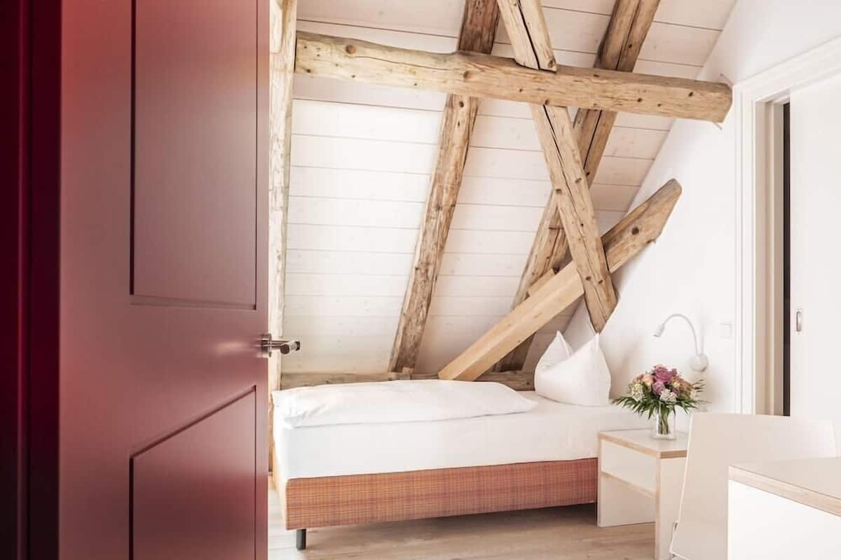 A bedroom with a sloped ceiling, wooden beams, a bed, a bedside table, and a desk.
