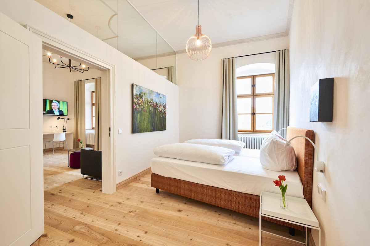 A modern bedroom with a wooden floor, a bed with white bedding, a small bedside table with a vase, a painting on the wall, and a television in the background.