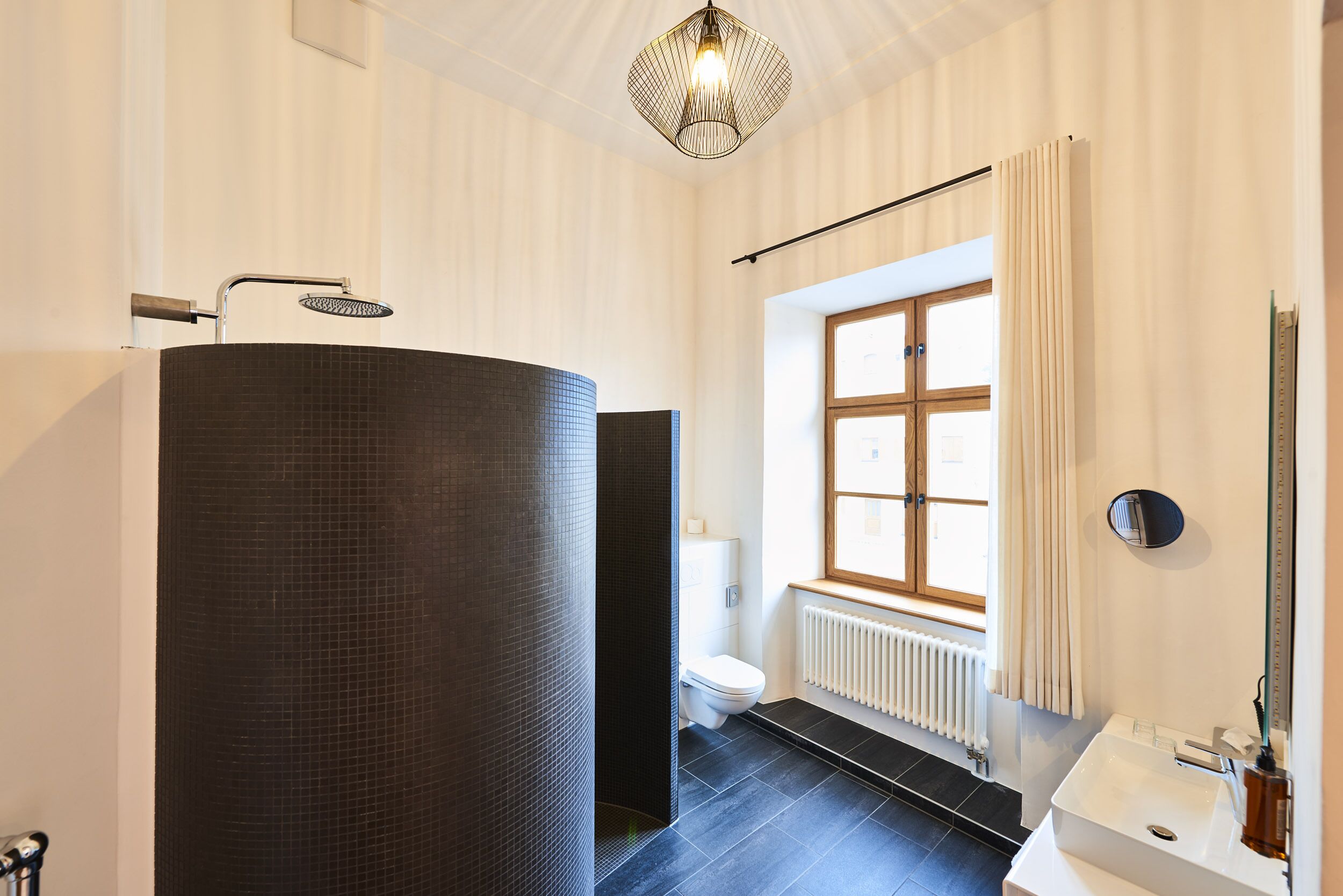 A modern bathroom with a large dark tiled sink, a white toilet, and a window with curtains.