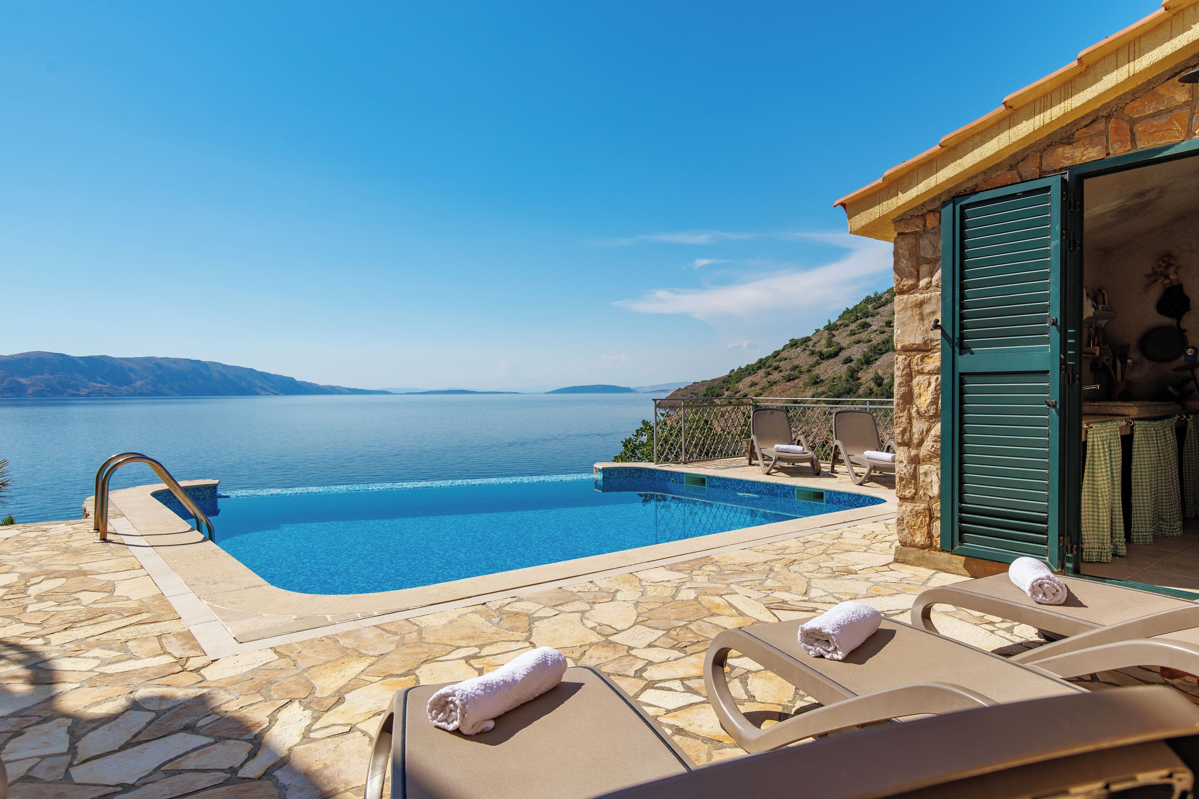 Una piscina con vistas al mar, suelo de piedra y sillones de descanso con toallas.