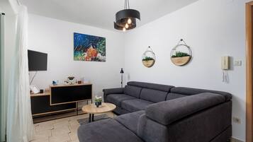 Uma sala de estar moderna com um sofá modular, uma televisão de tela plana, um quadro e dois vasos de plantas fixados na parede.