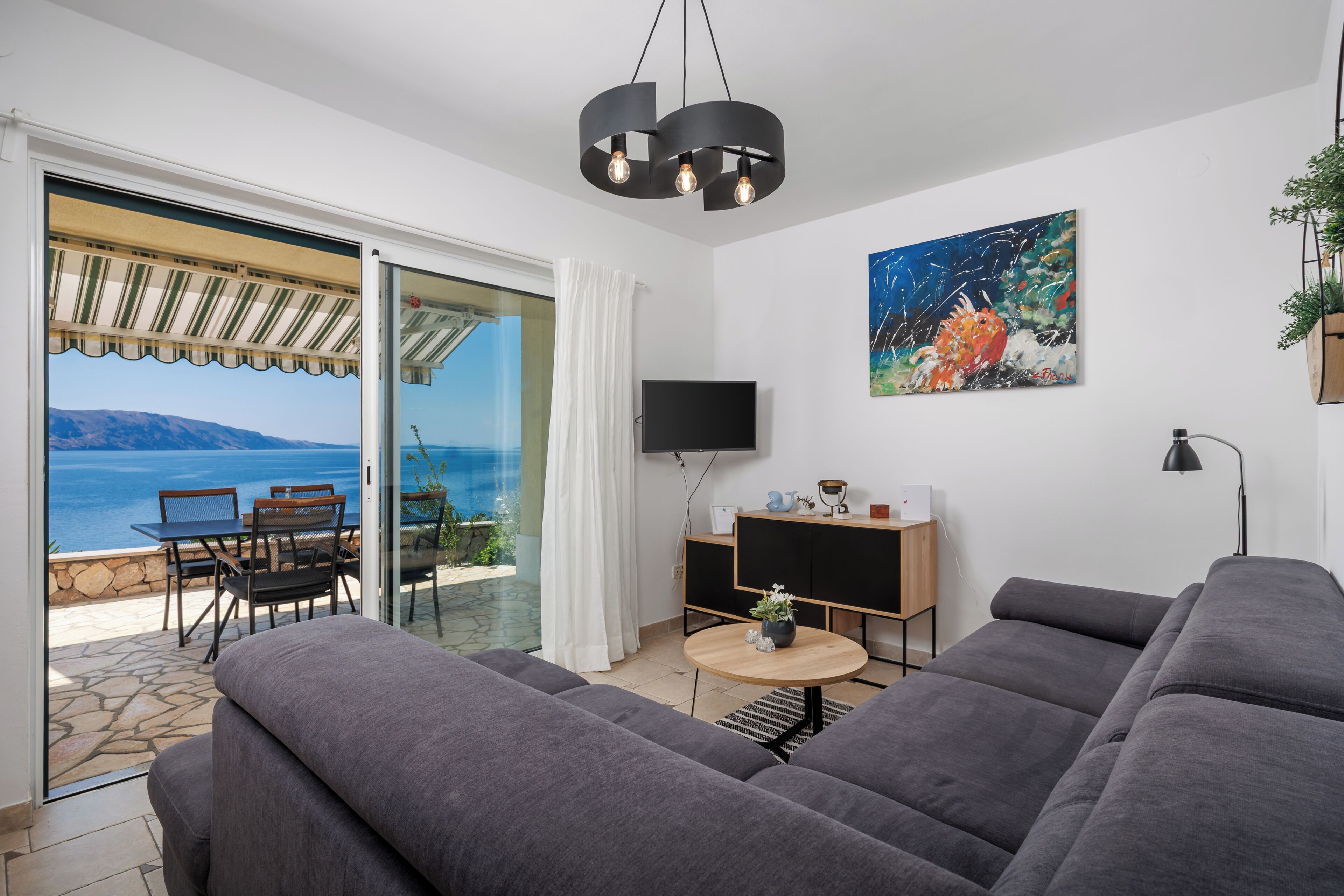 Un salon moderne avec un canapé gris, une petite table ronde et un téléviseur posé sur un meuble en bois. Un tableau orne le mur, et une porte vitrée offre une vue sur la mer.