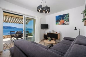 A modern living room with a grey sofa, a small round table, and a TV on a wooden cabinet. A painting is on the wall, and there's a view of the sea through a glass door. - Villa with pool and private waterfront (Jablanac)