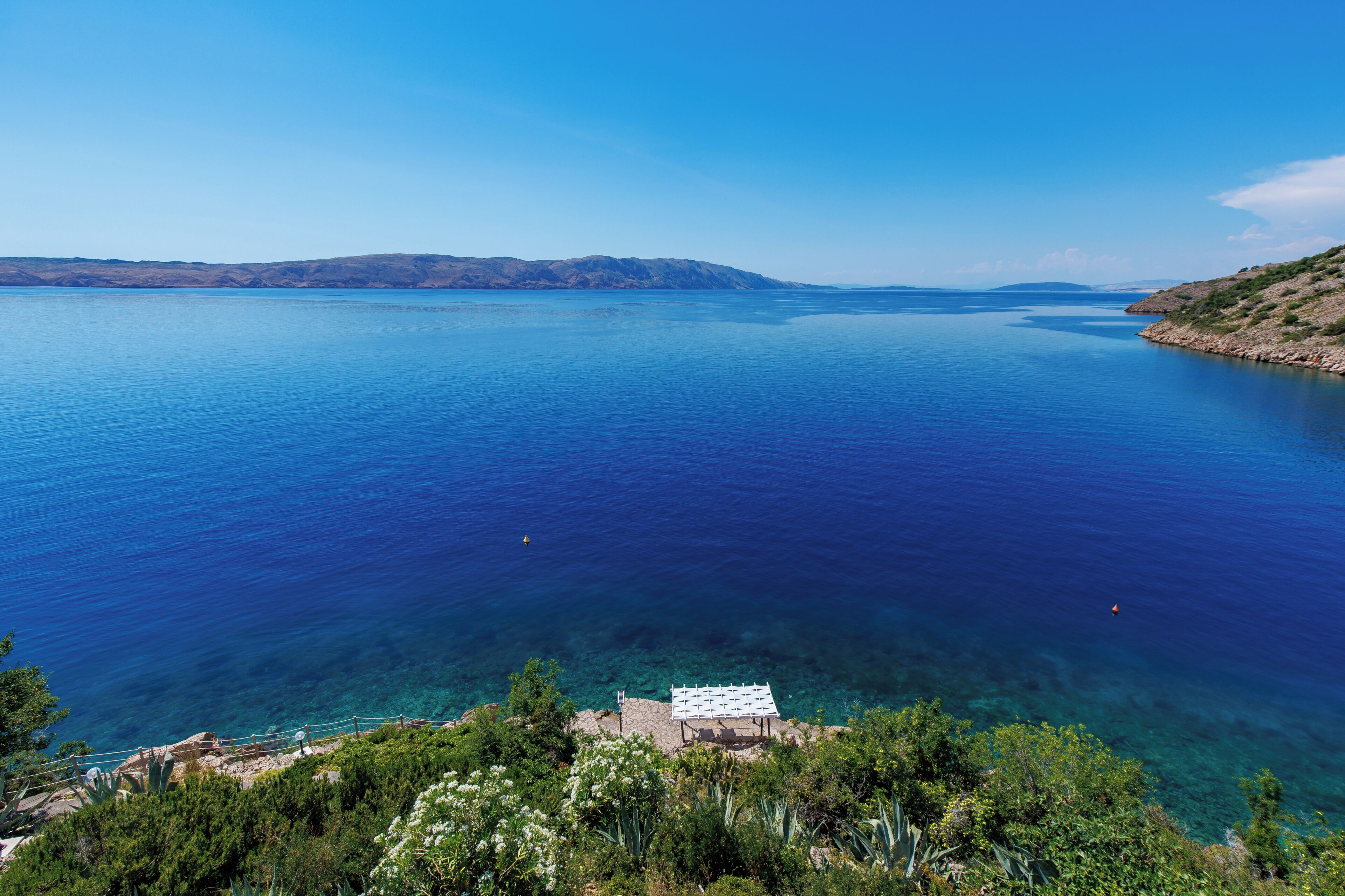 Vista sulla costa con mare blu cristallino, terreno roccioso e un edificio bianco con tetto.