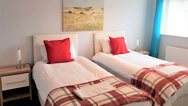 Quarto de hotel com duas camas, cada uma com roupa de cama branca e almofadas vermelhas, um quadro na parede, uma mesa de cabeceira com abajur e uma janela com cortinas azuis.