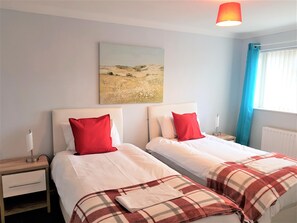 A hotel room with two beds, each with white bedding and red pillows, a painting on the wall, a bedside table with a lamp, and a window with blue curtains. - Penllech House - Huku Kwetu (Nottingham)