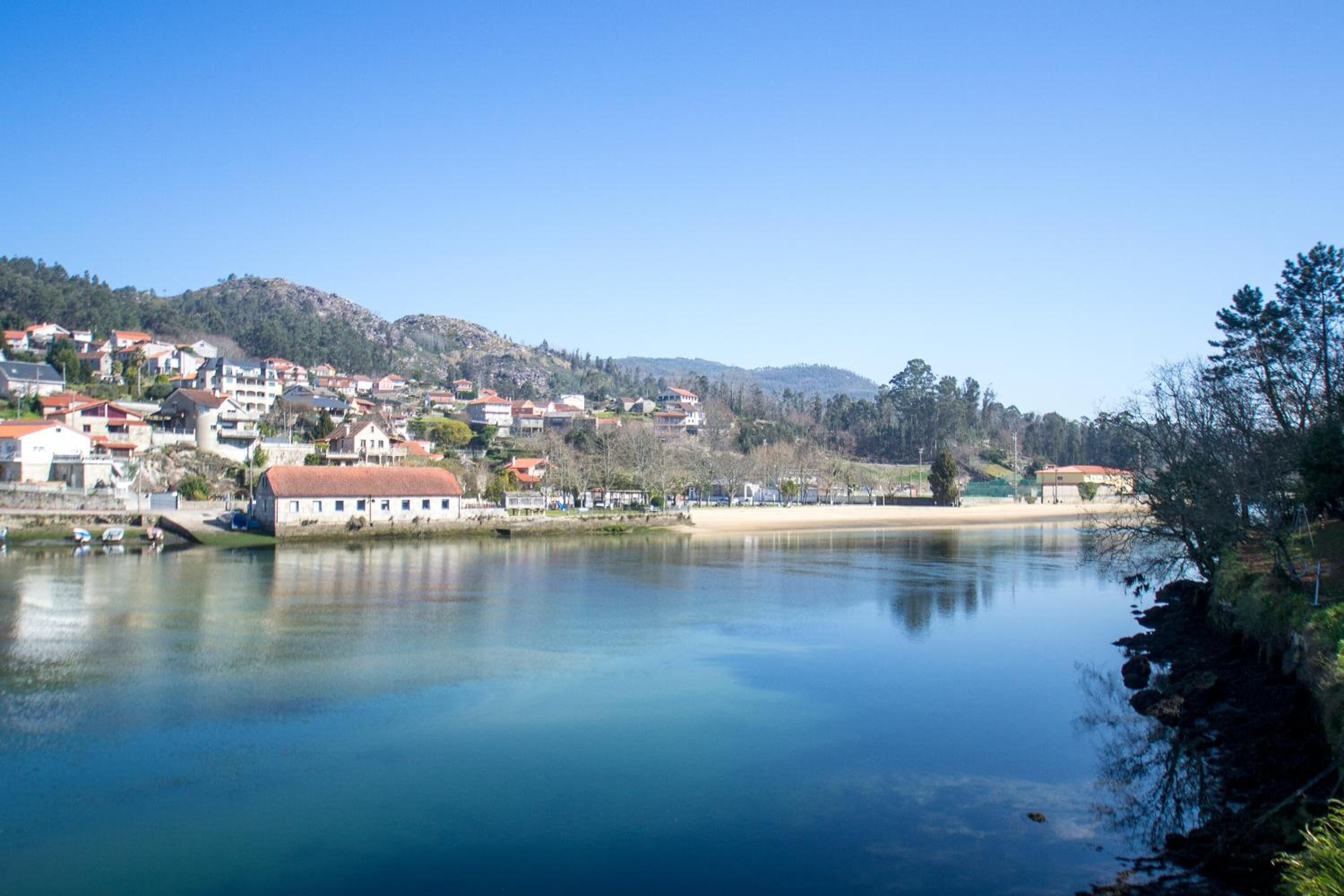 Un pueblo costero con edificios a lo largo del agua, una playa de arena y colinas al fondo.