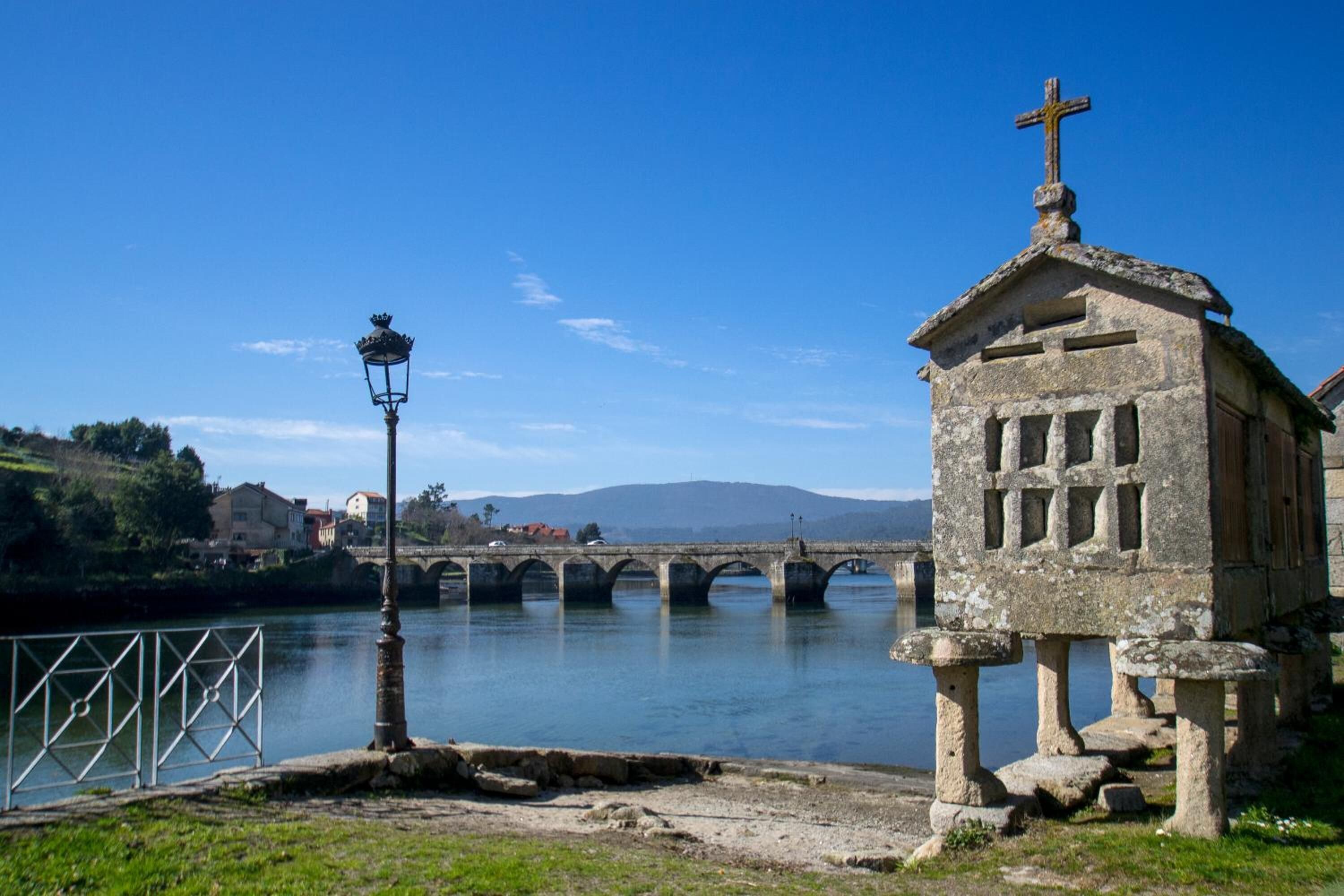 A stone chapel with a cross on top, a bridge in the background, and a lamp post in the foreground.