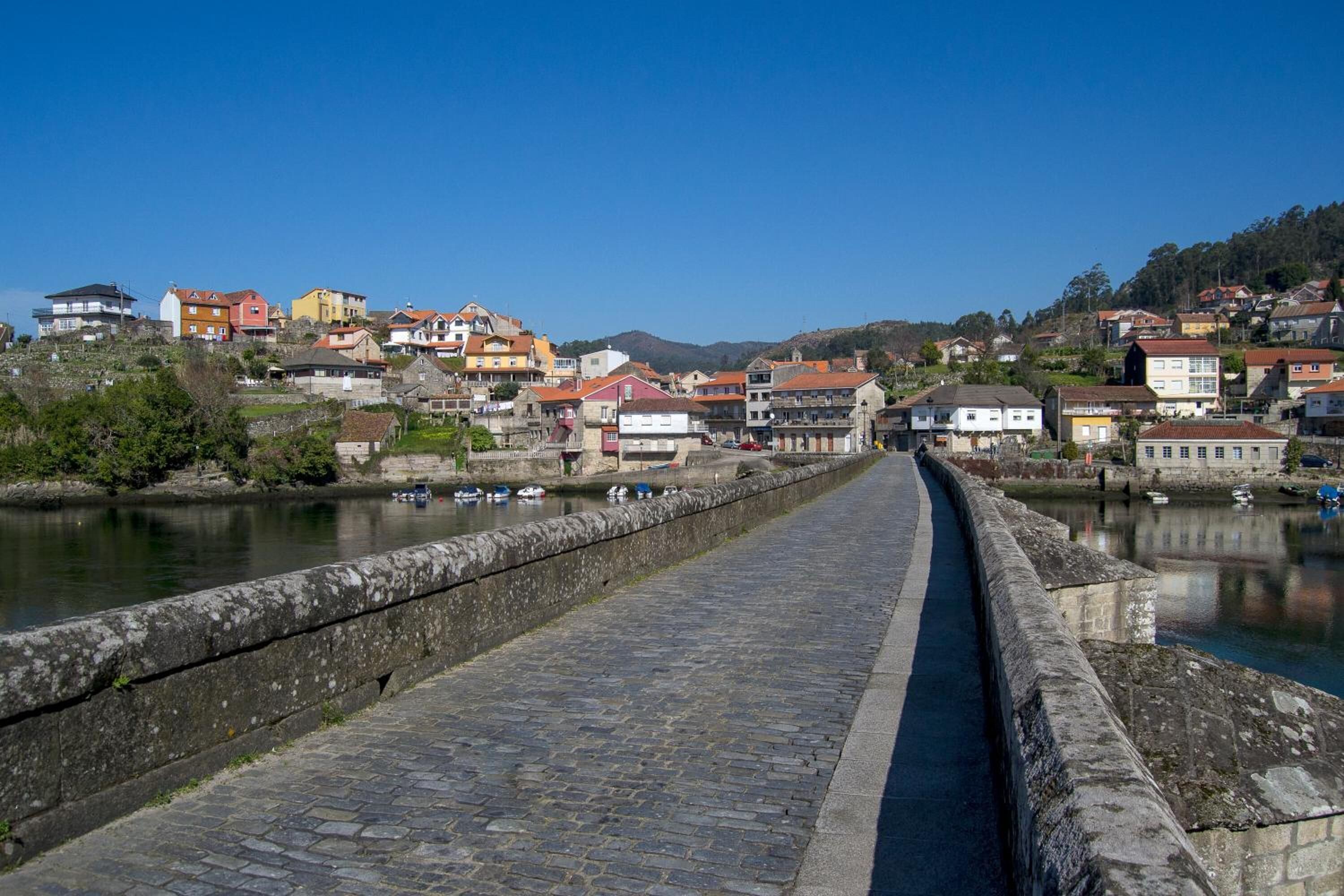 A stone bridge leading to a town with colorful buildings and a river.