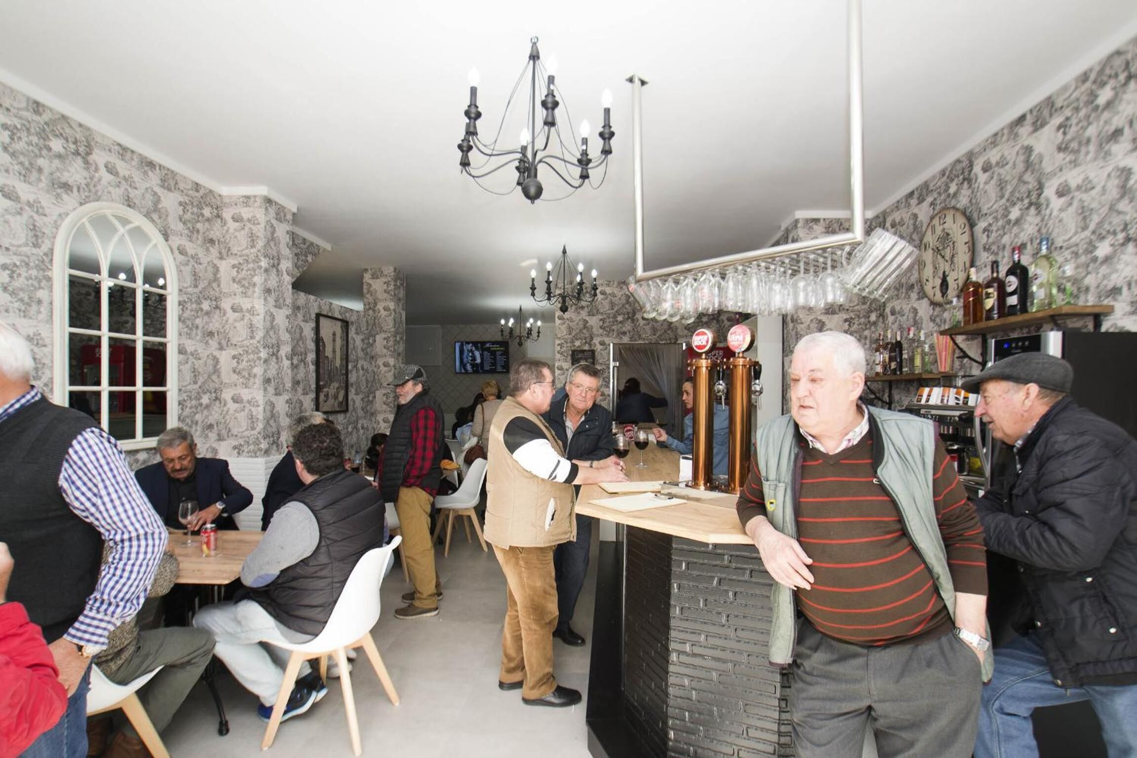 Reunión social en una sala con papel tapiz estampado, un candelabro y una zona de bar con vasos y botellas.