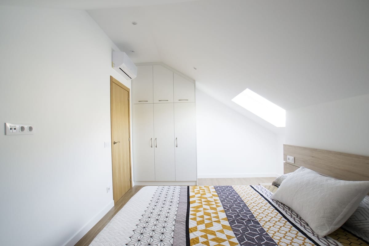 Ein Schlafzimmer mit schrägem Dach, einem Oberlicht, einem Bett mit gemusterten Betttextilien und einem Kleiderschrank.