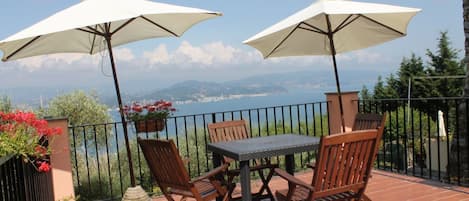 A terrace with a table and chairs, two white umbrellas, and a view of the sea and mountains.