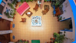 Une pièce avec un sol carrelé, une mosaïque décorative au centre, un canapé rouge à motifs, des chaises en bois, un vélo bleu et diverses plantes en pot.