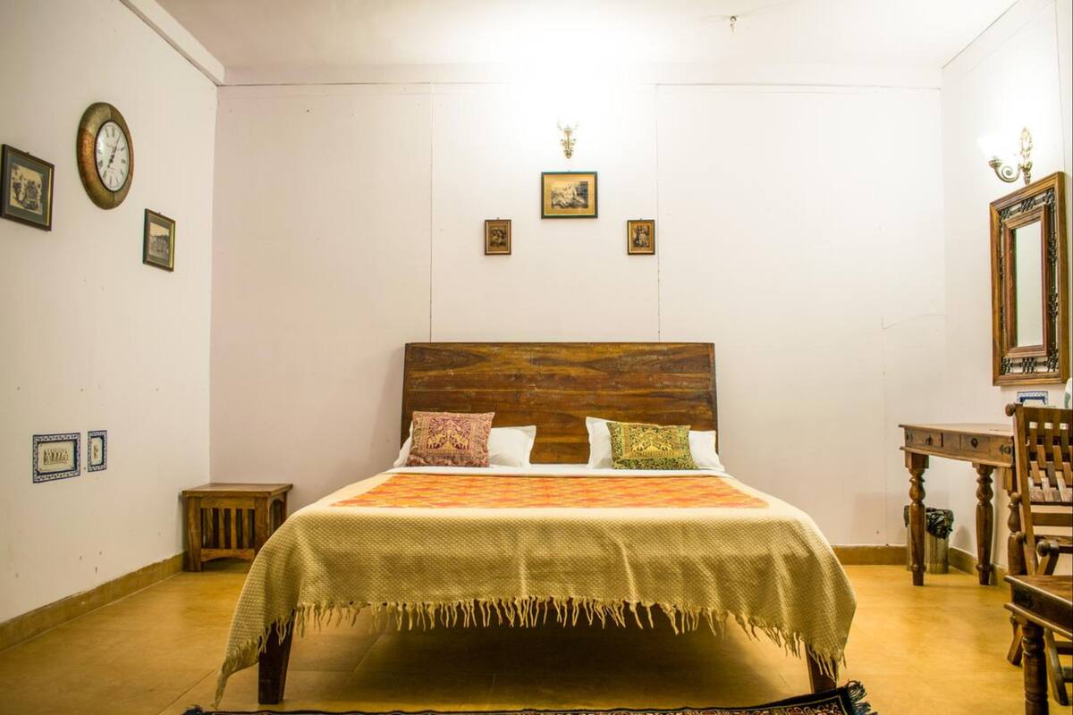 Een slaapkamer met een houten hoofdbord, een bed met een bedovertrek met patroon, een klein nachtkastje, een stoel en een spiegel aan de muur.